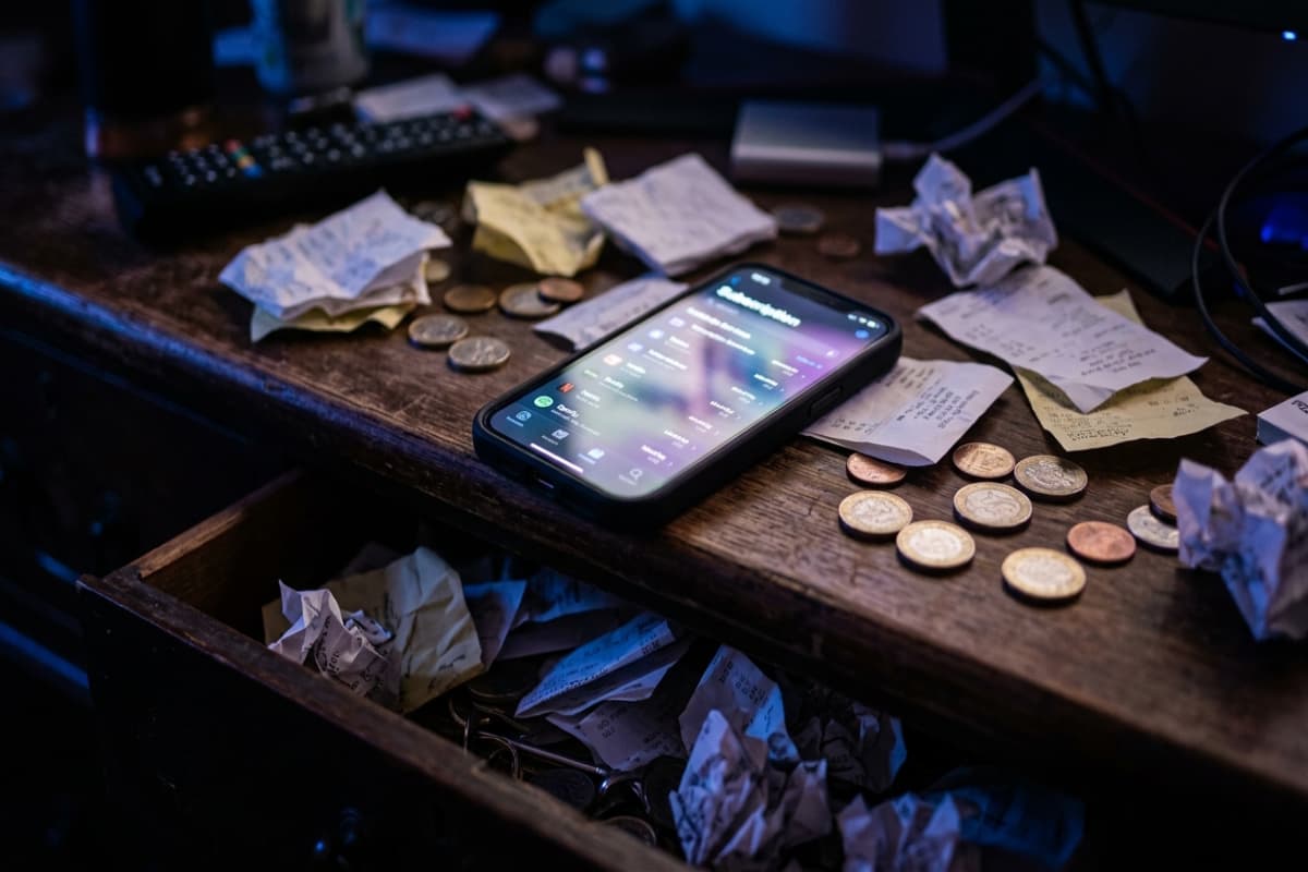 Smartphone showing a subscription list glowing on a dark desk, surrounded by scattered receipts and a drawer of more receipts
