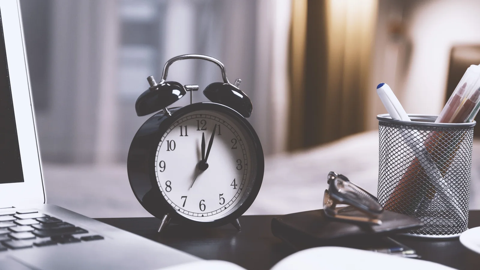 Black twin-bell alarm clock on a desk beside a laptop and pencil holder