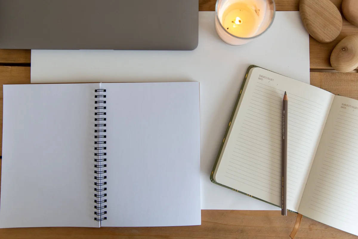 Blank notebooks and pencil beside part of a closed laptop and candle on a wooden desk